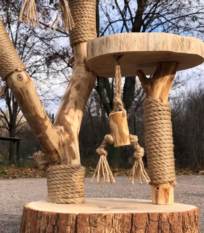 May include: A natural wood cat tree with a round base and a circular platform. The tree features multiple branches wrapped in thick rope, with a hanging wooden toy. The cat tree is designed for indoor use.
