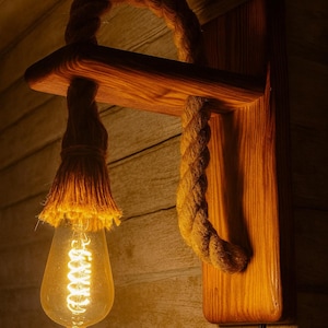 Op de afbeelding: Een rustieke wandlamp met een houten basis en plank, met een decoratief touwontwerp. De lamp heeft een Edison-lamp in vintage-stijl. De lamp is op een houten muur gemonteerd.