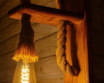 May include: A wall-mounted light fixture featuring a natural wood bracket and a thick, tan jute rope that loops over the wooden arm. A clear glass Edison-style light bulb with a visible glowing filament hangs from the end of the rope.