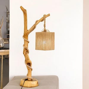 Può includere: Una lampada da tavolo con base in ramo di legno marrone chiaro naturale e supporto a fetta di legno circolare. Una spessa corda beige è avvolta attorno al ramo e sostiene un paralume quadrato rivestito di sottili fibre beige verticali.
