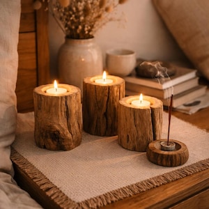 May include: Three wooden candle holders with lit candles on a beige placemat. The candle holders are made from sections of tree trunks. A small wooden incense holder with a lit incense stick is also present. A vase with dried flowers is in the background.
