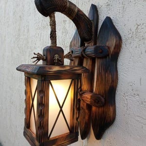 Op de afbeelding: Een wandlamp gemaakt van donker, geschroeid hout met een natuurlijke houtnerf. De vierkante lamp heeft matglazen panelen en hangt aan een dik, geknoopt touw aan een gebogen houten arm die is bevestigd aan een verticaal achterpaneel.