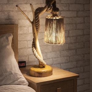 May include: A distinctive bedside lamp with a natural aesthetic. The lamp has a wooden base and a branch-shaped frame entwined with rope. The lampshade is cylindrical and made of fringed, natural-toned material. The lamp is on a wooden bedside table.