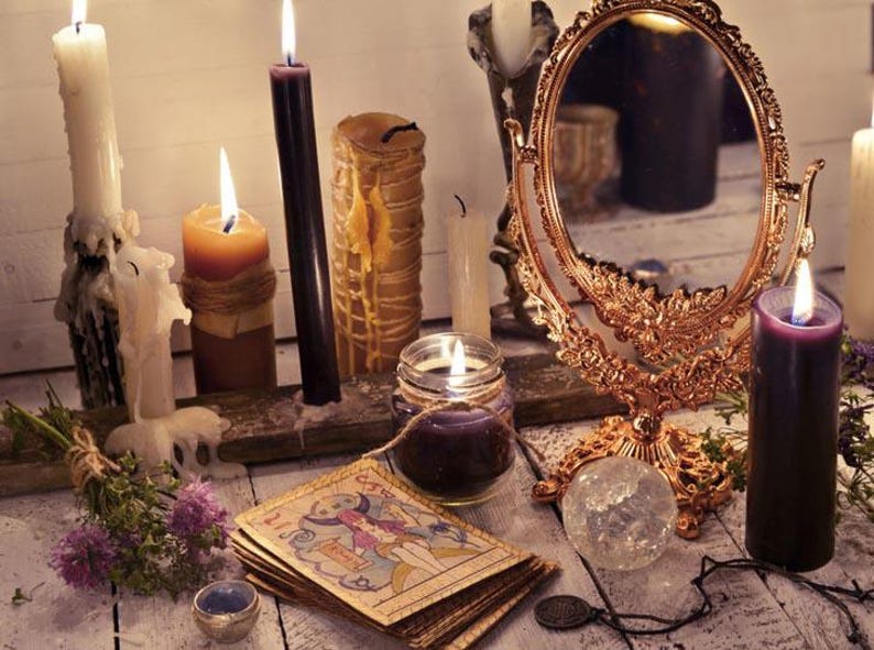 May include: A collection of candles, a mirror, and tarot cards arranged on a wooden surface. The candles are various colors, including white, black, and brown. The mirror is ornate and has a gold frame. The tarot cards are spread out on the surface, and some of them are visible. The scene is reminiscent of a mystical or spiritual setting.