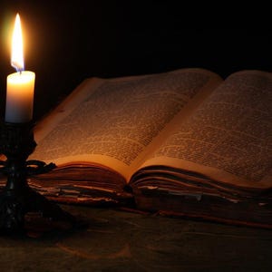 May include: A lit candle on a candlestick illuminates an open antique book with yellowed pages. The book is lying on a dark surface.