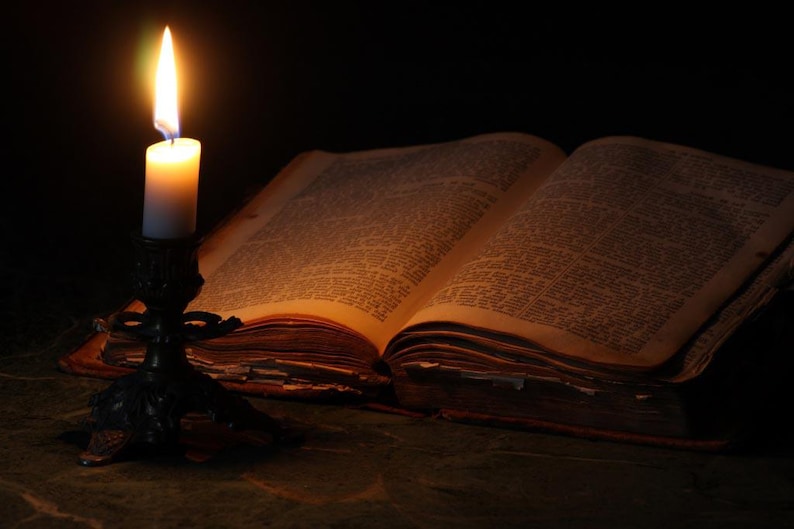 May include: A lit candle on a candlestick illuminates an open antique book with yellowed pages. The book is lying on a dark surface.