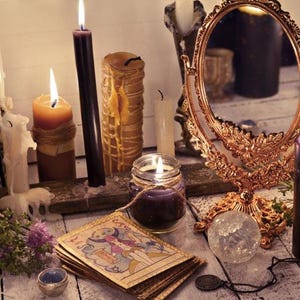 May include: A collection of candles, a mirror, and tarot cards arranged on a wooden surface. The candles are various colors, including white, black, and brown. The mirror is ornate and has a gold frame. The tarot cards are spread out on the surface, and some of them are face up.