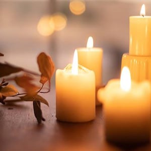 May include: A group of five candles burning brightly in a warm, inviting setting. The candles are all different sizes and shapes, and they are arranged in a cluster on a wooden surface. The candles are surrounded by a few dried leaves, adding to the cozy ambiance.