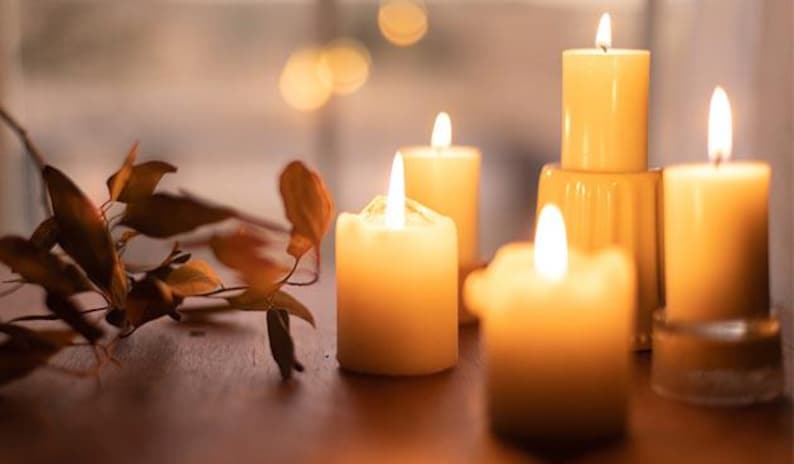 May include: A group of five candles burning brightly in a warm, inviting setting. The candles are all different sizes and shapes, and they are arranged in a cluster on a wooden surface. The candles are surrounded by a few dried leaves, adding to the cozy ambiance.