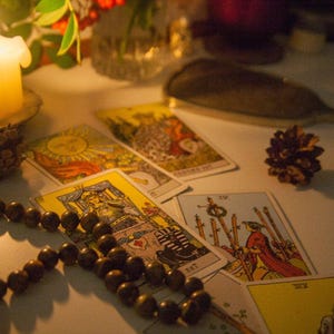 May include: A lit candle sits on a table with tarot cards spread out around it. A wooden beaded necklace is also on the table. The cards are from a traditional tarot deck and feature illustrations of people and symbols.