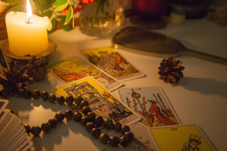 May include: A lit candle sits on a table with tarot cards spread out around it. A wooden beaded necklace is also on the table. The cards are from a traditional tarot deck and feature illustrations of people and symbols.