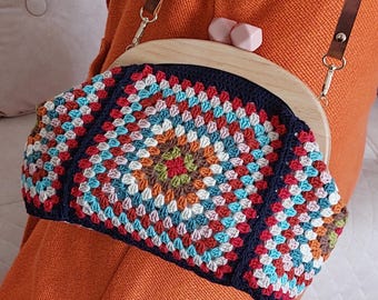 Puede incluir: Un bolso de ganchillo cuadrado de la abuela colorido con un marco de madera y una correa de cuero marrón. El bolso lo lleva una persona que lleva un suéter naranja.