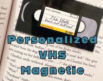 May include: A black VHS tape bookmark with the text "The State Beavis and Butthead" and "E-180" printed on it. The bookmark is sitting on top of a book.