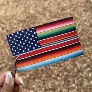 May include: A rectangular sticker featuring a unique design that combines the American flag with a colorful serape pattern. The flag's stars and stripes are visible, with the serape adding vibrant stripes of red, orange, green, and blue.