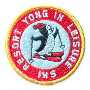 Könnte beinhalten: Runder, gestickter Aufnäher mit einem Ski fahrenden Cartoon-Pinguin. Der Aufnäher hat einen roten Rand mit gelben Nähten. Der Text lautet "YONG IN LEISURE SKI RESORT". Der Pinguin ist schwarz-weiß und trägt einen roten Hut und Schal.