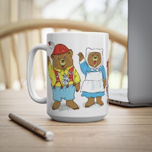 May include: White ceramic mug with a colorful illustration of three bears wearing different costumes. The bears are wearing a red and white hat, a white dress with a blue apron, and a pink dress. The mug has a handle on the left side.