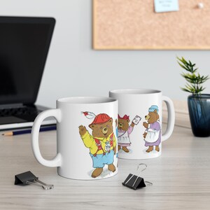 May include: Two white ceramic mugs with illustrations of bears. One mug features a bear wearing a red construction hat and holding a feather. The other mug features three bears, one wearing a blue dress and holding a piece of paper.
