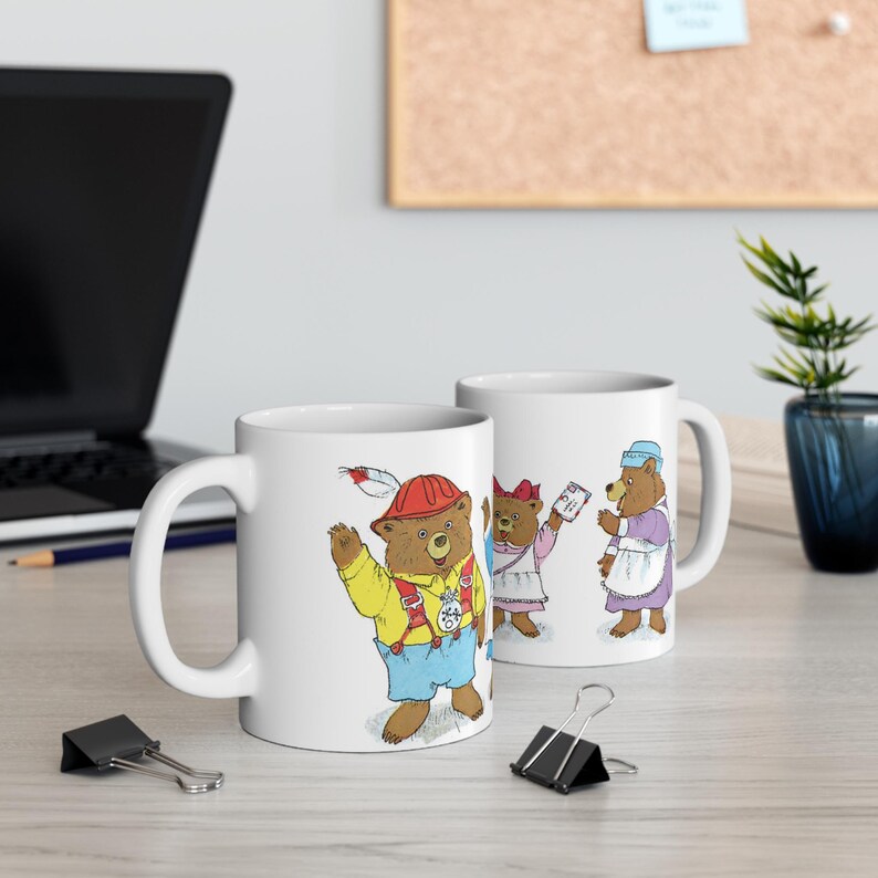 May include: Two white ceramic mugs with illustrations of bears. One mug features a bear wearing a red construction hat and holding a feather. The other mug features three bears, one wearing a blue dress and holding a piece of paper.