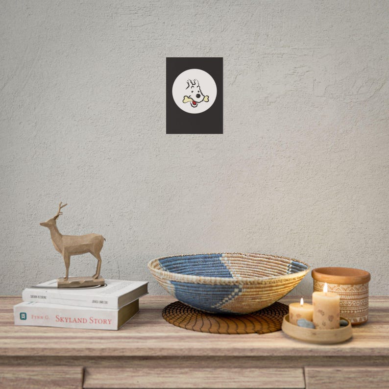 Puede incluir: Un estampado enmarcado que presenta un perro de dibujos animados con un hueso en la boca, sobre un fondo negro dentro de un c&iacute;rculo blanco. El estampado se exhibe sobre una mesa de madera con elementos decorativos, como una figura de ciervo y un cuenco tejido.