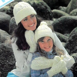 May include: A mother and daughter wearing matching white knitted hats and mittens. The hats have a cable knit pattern and the mittens have a leaf design.