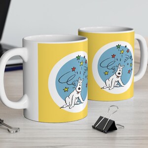 May include: Two white ceramic mugs with yellow backgrounds and blue circles featuring a cartoon white dog with a red tongue and black eyes. The dog is surrounded by colorful stars. The text "Cleo" is written in black on the mugs.