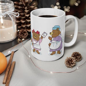 May include: White ceramic mug with a colorful illustration of two bears wearing dresses and holding a letter. The bear on the left is wearing a pink dress with a red bow and the bear on the right is wearing a blue dress with a white apron.