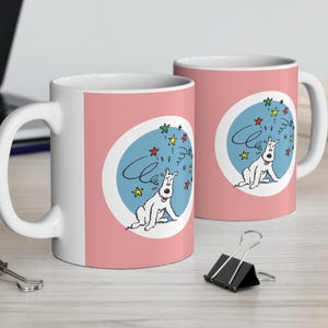 May include: Two white ceramic mugs with a pink background. Each mug features a cartoon dog inside a blue oval with colorful stars and the word "Cheers". The mugs are on a light wood surface.
