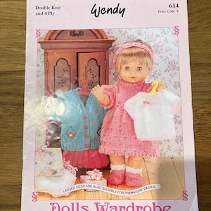 Könnte beinhalten: Ein Wendy 614 Strickmusterheft für eine Puppengarderobe mit 13 kompletten Outfits für eine Babypuppe. Das Cover zeigt eine Puppe in einem rosa gestrickten Kleid, Stirnband und Schühchen, die ein weißes Oberteil hält, daneben eine blaue Strickjacke.