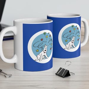 May include: Two white ceramic mugs with blue handles. Both mugs feature a blue background with a white circle containing a cartoon dog with stars around it. The text "Cléo" is written in a cursive font on the mug.