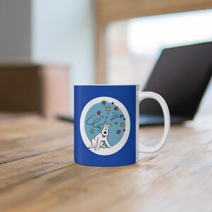 May include: A blue and white ceramic mug featuring a cartoon dog with a thought bubble and stars. The dog is white with black details, sitting inside a light blue circle. The mug has a white handle and is on a wooden surface.