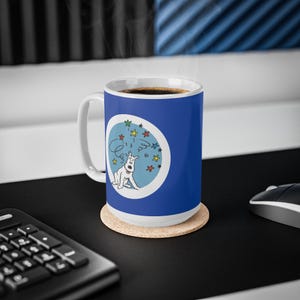 May include: A blue ceramic coffee mug with a white handle and a cartoon dog illustration. The mug features a white dog with a red collar inside a blue circle with stars. The mug sits on a cork coaster.