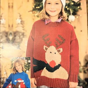 May include: A long-sleeved red knit sweater featuring a large reindeer graphic with brown antlers, a beige face, and a bright red nose, accented by a dark scarf. The sweater has a ribbed crew neckline, cuffs, and hem, worn over a plaid collared shirt.