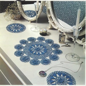 Può includere: Una collezione di centrini blu all'uncinetto di varie dimensioni, disposti su una superficie bianca. I centrini presentano intricati motivi floreali. Sono presenti anche oggetti decorativi come specchi, bottiglie di profumo e candelabri, creando un'estetica vintage.