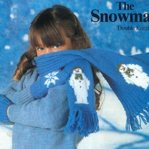 May include: A blue knitted scarf and mittens set with snowman and snowflake patterns. The scarf has a fringed edge. The text "The Snowman Double Knitting" is visible in the background. The mittens and scarf are a matching set.
