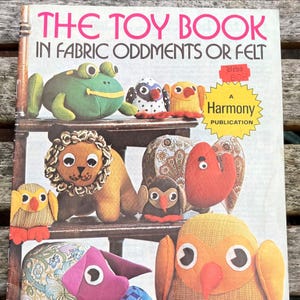 May include: A book cover titled THE TOY BOOK IN FABRIC ODDMENTS OR FELT featuring various handmade stuffed animal toys including a green frog, a lion, an elephant, and several birds. A yellow starburst label reads A HARMONY PUBLICATION.