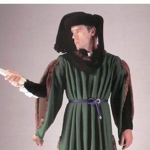 Puede incluir: Un disfraz medieval verde con un sombrero y capa negros, ribeteado con piel marrón. El disfraz presenta un cinturón morado y puños blancos. La persona sostiene un pergamino blanco.