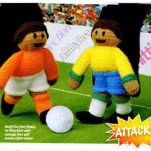 May include: Two knitted football player dolls on a green pitch, one orange and one yellow, with a white ball. The orange doll wears white shorts and orange socks. The yellow doll wears blue shorts. A yellow and orange starburst graphic says "ATTACK!"