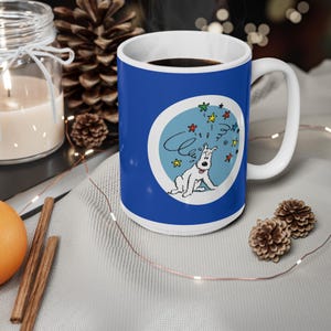 May include: A white ceramic mug with a blue exterior and a white handle. The mug features a cartoon illustration of a white dog with stars and the word "C" inside a blue circle.