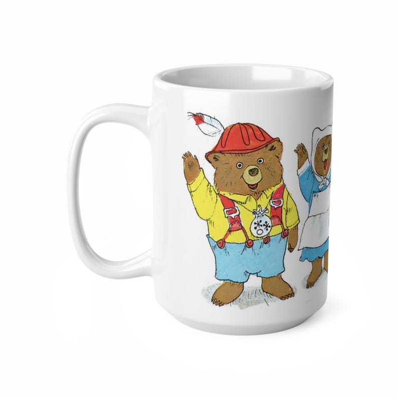 May include: White ceramic mug with a colorful illustration of three bears. The bear in the center is wearing a red hat and a yellow shirt with suspenders. The bear on the left is wearing a blue dress and the bear on the right is wearing a red dress.