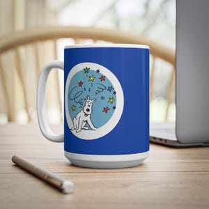 May include: A white ceramic mug with a blue background and a cartoon illustration of a white dog with a swirl and star design. The mug has a white handle and base, and is sitting on a wooden surface.