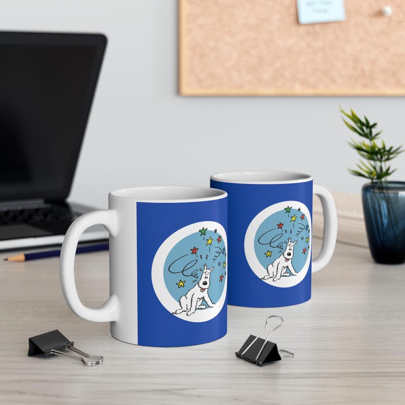 May include: Two white ceramic mugs with a blue background. Each mug features a cartoon dog design inside a light blue oval, with colorful stars and the word "Cheers". The mugs are on a light-colored surface.