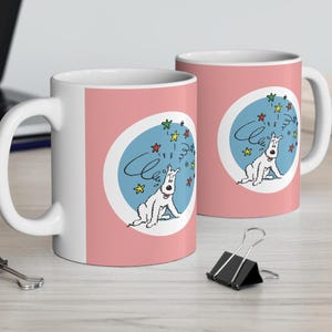 May include: Two white ceramic mugs with pink backgrounds and a cartoon dog design. The dog is white with black eyes and a red collar. The dog is surrounded by blue stars and the word "Cleo" is written in a cursive font.