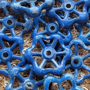 Puede incluir: Un conjunto de manijas de válvula azules en forma de estrella con un acabado desgastado. Las manijas, con un orificio central, están dispuestas sobre un lecho de material similar a la paja. La pintura azul muestra signos de desgaste, dándoles un aspecto vintage.