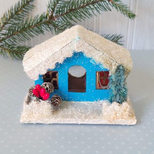 Puede incluir: Una casa azul en miniatura con techo y base cubiertos de nieve, adornada con piñas y bayas rojas. La casa presenta ventanas rectangulares y arqueadas, y un pequeño árbol verde. Una ramita de hoja perenne está en el fondo.