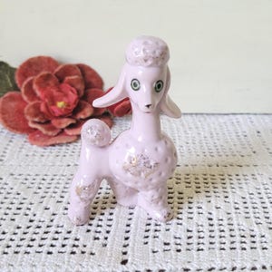 May include: A light pink ceramic poodle figurine featuring textured fur details and subtle gold-toned accents on the head, chest, and legs. The dog has green eyes, a black nose, and stands in a classic pose with its head turned slightly.