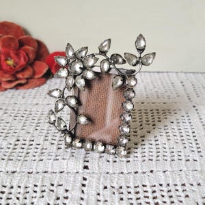 May include: A small rectangular picture frame featuring a dark metal finish and clear, faceted crystal-like stones arranged in a floral and vine pattern along the side and top edges. The center of the frame contains a plain, light brown insert.