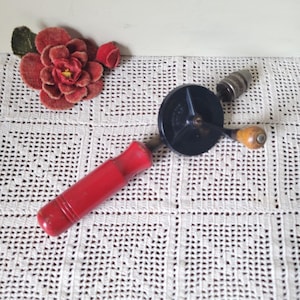 May include: A vintage hand drill featuring a bright red cylindrical handle, a black circular gear housing, a small wooden crank handle, and a metal chuck. The tool is positioned diagonally across the frame, showcasing its mechanical components.