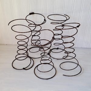 May include: A collection of vintage, rusty metal springs, each with a coiled design and circular top and bottom. The springs vary in size and shape, with a weathered, aged appearance. The background is a white wooden surface.