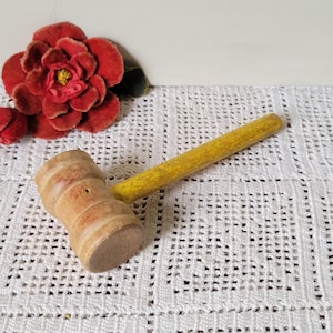 Puede incluir: Un mazo de madera con mango amarillo y cabeza marrón claro. El mazo está sobre una superficie de encaje blanco. Una flor roja está en el fondo.
