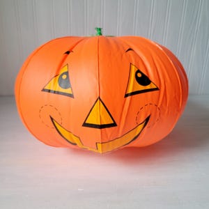 Puede incluir: Una calabaza inflable naranja con una cara de jack-o'-lantern. La cara presenta ojos triangulares, una nariz triangular y una boca curva. La calabaza tiene un tallo verde y está diseñada para Halloween.